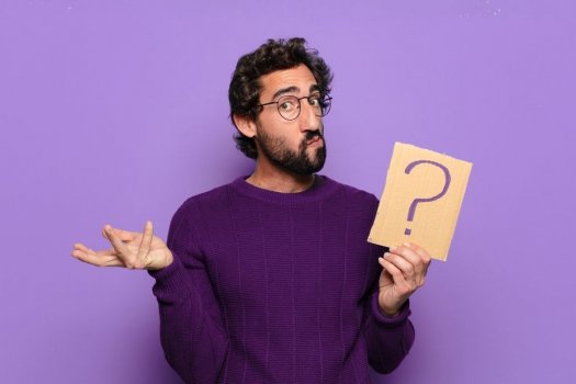 Ein Mann im lila Pullover der ein Fragezeichen auf Pappe in der Hand hält und ratlos guckt.