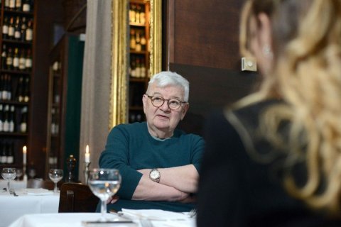 Joschka Fischer, ehemaliger Außenminister und Vizekanzler der Bundesrepublik Deutschland im Gespräch, Foto: E.ON Stiftung Das Bild zeigt ein Restaurant mit zwei Personen, die an einem Tisch sitzen. Die Person im Vordergrund hat langes, gewelltes blondes Haar und ist von der Kamera abgewandt, so dass nur der Hinterkopf zu sehen ist. Die Person im Hintergrund, deren Gesicht verschwommen ist, hat kurze weiße Haare und trägt einen blaugrünen Pullover. Sie sitzen mit verschränkten Armen und tragen eine Uhr am linken Handgelenk. Der Tisch ist mit weißen Tischdecken, Gläsern und Besteck gedeckt. Im Hintergrund ist eine Wand mit Regalen voller Weinflaschen zu sehen, und auf dem Tisch ist eine brennende Kerze zu sehen. Die Umgebung deutet auf eine formelle Essumgebung hin.