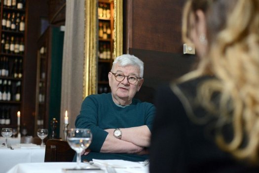 Das Bild zeigt ein Restaurant mit zwei Personen, die an einem Tisch sitzen. Die Person im Vordergrund hat langes, gewelltes blondes Haar und ist von der Kamera abgewandt, so dass nur der Hinterkopf zu sehen ist. Die Person im Hintergrund, deren Gesicht verschwommen ist, hat kurze weiße Haare und trägt einen blaugrünen Pullover. Sie sitzen mit verschränkten Armen und tragen eine Uhr am linken Handgelenk. Der Tisch ist mit weißen Tischdecken, Gläsern und Besteck gedeckt. Im Hintergrund ist eine Wand mit Regalen voller Weinflaschen zu sehen, und auf dem Tisch ist eine brennende Kerze zu sehen. Die Umgebung deutet auf eine formelle Essumgebung hin.