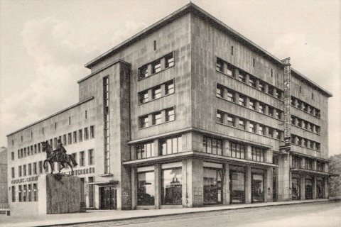 Das Bild zeigt ein großes, mehrstöckiges Gebäude im Stil der Neuen Sachlichkeit. Die Fassade ist schlicht und geometrisch gestaltet, mit vielen rechteckigen Fenstern in gleichmäßigen Reihen. An der Ecke des Gebäudes ist ein senkrechtes Schild mit der Aufschrift „LICHTBURG“ angebracht. Im Vordergrund steht eine Reiterstatue auf einem hohen Sockel. Das Gebäude wirkt monumental und funktional, mit großen Schaufenstern im Erdgeschoss. Es handelt sich um die historische Lichtburg in Essen, ein bekanntes Kino- und Veranstaltungsgebäude.