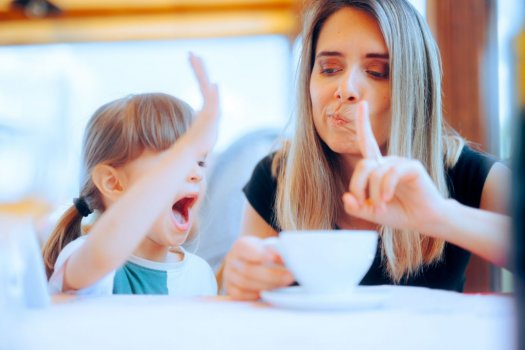 Eine junge Frau schaut skeptisch zu einem kleinen Mädchen, das neben ihr sitzt, und erhebt mahnend den Zeigefinger, während das Mädchen im Begriff ist, mit seiner rechten Hand auf eine weiße Tasse zu schlagen, die vor den beiden auf einem Tisch steht. Der ausgestreckte Arm verdeckt in dem Moment die Augen des Mädchens, während sein Mund vor Übermut weit geöffnet ist. 
