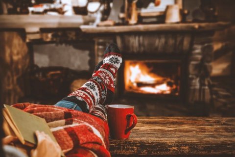Foto: S_Photo / shutterstock Links vorne im Bild erkennen wir zwei Beine in einer Jeans und mit dicken gestrickten Wollsocken, die zum Teil von einer Wolldecke bedeckt sind. Zwei Hände halten ein Buch. Rechts neben den Beinen steht eine rote Tasse. Die Füße sind auf einer Holzbank abgelegt und im Hintergrund brennt ein Feuer in einem Kamin. Im Foto dominieren warme Farben wie Rot, Orange und Braun.