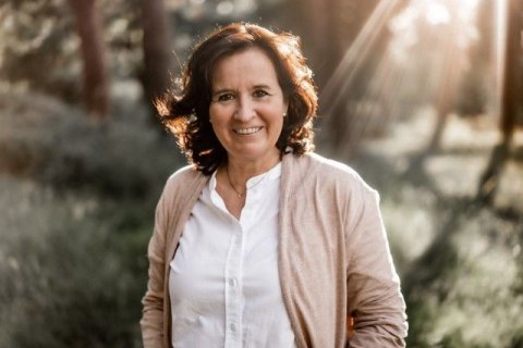 Die Dozentin Frauke Stoermer, Foto: privat Frau mit braunen Haaren, weißer Bluse und Strickjacke, die glücklich zu sein scheint, auf Waldlichtung; Sonnenlicht fällt seitlich von hinten ein und scheint durch die Haare