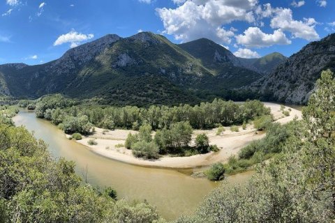 So sieht er aus, der stille Norden Griechenlands. Foto: Lothar Hoppen Das Bild zeigt eine Landschaft mit einem Fluss, der sich durch ein von Bergen umgebenes Tal schlängelt. Der Fluss fließt von der linken Seite des Bildes und macht eine deutliche Biegung nach rechts, wodurch entlang seiner inneren Kurve eine sandige Bank entsteht. Die sandige Fläche ist mit Gruppen von Bäumen und Sträuchern besiedelt, was auf eine üppige Uferzone hinweist. Die Berge im Hintergrund sind markant, mit steilen Hängen und felsigen Oberflächen, die mit Vegetationsflecken bedeckt sind. Das Tal ist dicht bewaldet, mit Bäumen, die sich vom Flussufer bis zum Fuß der Berge erstrecken. Der Himmel über mir ist klar mit vereinzelten Wolken und bildet eine Kulisse für das bergige Gelände.