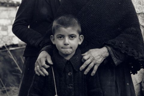 Der junge Argyris, Foto: Fontana Film Das Bild zeigt drei Individuen. Im Vordergrund ist ein Kind zu sehen. Das Kind trägt ein zugeknöpftes Kleidungsstück mit sichtbarem Kragen. Hinter dem Kind sind zwei Erwachsene anwesend. Der Erwachsene auf der linken Seite trägt ein dunkles, langärmeliges Kleidungsstück, der linke Arm ruht auf der Schulter des Kindes. Der Erwachsene auf der rechten Seite trägt einen strukturierten Schal oder ein Tuch, die Hände ruhen auf den Schultern des Kindes. Die Hände des Erwachsenen auf der rechten Seite sind deutlich zu sehen und zeigen Altersspuren. Im Hintergrund gibt es eine Steinmauer und einige undeutliche Elemente.