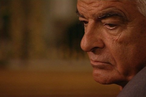 Argyris Sfountouris im Alter von 66 Jahren, Foto: Fontana Film Das Bild zeigt ein verschwommenes Gesicht auf der rechten Seite, während der Rest des Hintergrunds unscharf ist. Der Hintergrund scheint eine Inneneinrichtung mit undeutlichen Formen und Farben zu sein, die möglicherweise auf Objekte oder Dekorationen hinweisen, aber es sind keine spezifischen Details zu erkennen.