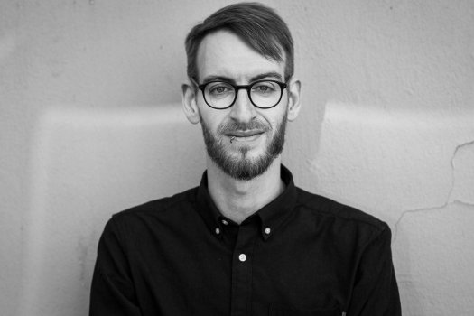Das Bild zeigt eine Person, die an einer strukturierten Wand steht. Die Person trägt ein dunkles Hemd mit sichtbaren Knöpfen am Kragen und auf der Vorderseite. Das Hemd scheint ordentlich abgetragen zu sein, der Kragen ist zugeknöpft. Das Haar der Person ist ordentlich gestylt, mit einem Seitenscheitel. Die Wand hinter der Person weist sichtbare Risse und eine raue Oberfläche auf, was darauf hindeutet, dass sie aus Gips oder einem ähnlichen Material besteht. Die Beleuchtung im Bild ist gleichmäßig und ermöglicht eine klare Sichtbarkeit des Hemdes und der Wanddetails.