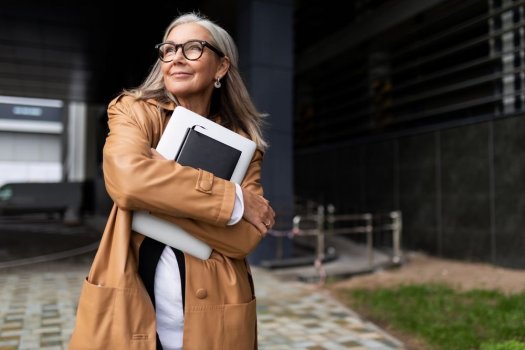 Eine Frau mit langen grauen Haaren, dunkler Brille, Lederjacke und Ohrringen steht draußen vor einem Bürogebäude. Sie hält ein Laptop und ein Notizbuch in ihren Armen vor der Brust und schaut optimistisch in die Ferne - weg vom Bürogebäude. 