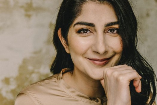 Das Bild zeigt eine Person mit langen, dunklen Haaren, die über die rechte Schulter fallen. Sie trägt eine glänzende, beige Bluse mit kleinem Stehkragen und Knopf am Ausschnitt. Eine Hand ist angewinkelt; die Finger ruhen locker unter dem Kinn. Die Person blickt direkt in die Kamera, die Lippen formen ein leises Lächeln. Der Hintergrund wirkt texturiert in creme- und ockerfarbenen Tönen.