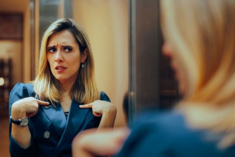 Foto: Nicoleta Ionescu / Shutterstock Eine Frau mit langen Haaren, Ohrring und einem Blazer betrachtet sich selbst zweifelnd im Spiegel. Wir sehen ihr Spiegelbild scharf und frontal, sie selbst nur unscharf von hinten. Ihre Stirn ist gekräuselt, der Blick unsicher und sie zeigt fragend mit beiden Zeigefingern auf sich selbst, als wollte sie sagen: "Kann ich das? Darf ich das? Bin ich das?"
