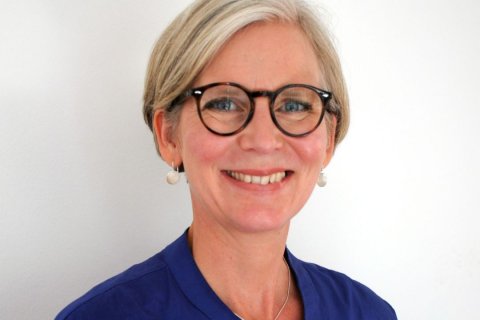 Die Philosophin Ina Schmidt, Foto: Melanie Zwiehoff-Bremicker Das Bild zeigt eine Frau mit kurz geschnittenem, hellblond-grauem Haar. Sie trägt eine runde, dunkle Brille und lächelt freundlich. Ihre blauen Augen sind gut sichtbar. Sie trägt kleine runde Ohrringe und ein Oberteil in kräftigem Blau mit schmalem Stehkragen. Der Hintergrund ist weiß und neutral.