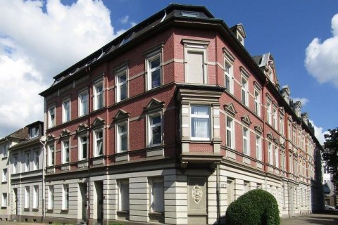 Häuserblock des Bauunternehmers H. Elting im Eltingviertel, Foto: Robert Welzel Das Bild zeigt ein großes, dreistöckiges Eckgebäude mit Mansardendach. Das Gebäude ist aus rotem Backstein gebaut und verfügt über dekorative Steinelemente an den Fenstern und entlang der Fassade. Die Fenster sind symmetrisch angeordnet, wobei jedes Stockwerk eine Reihe von rechteckigen Fenstern hat, die von Steinstürzen und Fensterbänken eingerahmt sind. Das Erdgeschoss hat eine hellere Steinfassade, die einen Kontrast zu den roten Ziegeln darüber bildet. Ein kleiner, abgerundeter Busch befindet sich an der Ecke des Gebäudes, angrenzend an den Bürgersteig. Die Architektur des Gebäudes erinnert an einen historischen Stil mit kunstvollen Details und einer prominenten Ecklage.