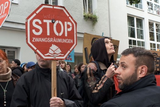 Das Bild zeigt eine Gruppe von Menschen, die an einem Protest teilnehmen. Im Mittelpunkt steht eine Person, die ein rotes achteckiges Schild in der Hand hält, das an ein Stoppschild erinnert und auf dem in weißer Schrift "STOP ZWANGSRÄUMUNGEN" steht. Das Schild wird auf einem Holzstab gehalten und ist prominent im Vordergrund positioniert.
Auf der linken Seite hält eine andere Person ein ähnliches, teilweise sichtbares Schild mit der gleichen Botschaft. Diese Person hat auffällig rote Haare und trägt einen Schal und eine Halskette.
Im Hintergrund sind weitere Protestschilder zu sehen, darunter eines aus Pappe mit dem Text "DIE STADT SIND WIR!". (Wir sind die Stadt!).
Der Protest findet vor einem Gebäude mit weißen Wänden und mehreren Fenstern statt. Auf einem Fenstersims über der Menschenmenge ist eine kleine Pflanze zu sehen. Die Szene ist überfüllt, Menschen stehen dicht an dicht, einige tragen Jacken und Kapuzenpullis.