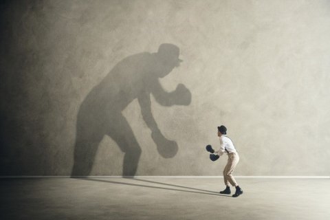 Foto: frankie / shutterstock Auf grauem Betonboden und vor einer großen grauen Betonwand ist klein eine Person von hinten zu sehen. Der Mensch trägt ein weißes Hemd, eine beigefarbene Hose und schwarze Schuhe, Hosenträger, einen Hut sowie Boxhandschuhe. Er ist zur Wand hin ausgerichtet und "bekämpft" seinen eigenen Schatten, der überlebensgroß an der Wand vor ihm "steht".