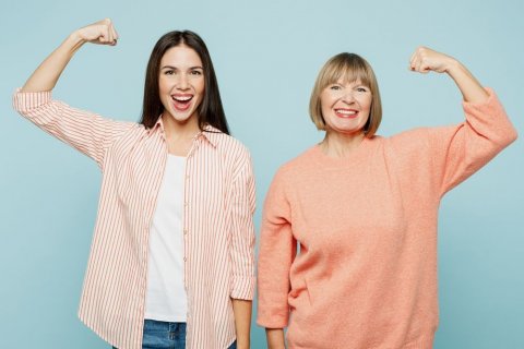 Foto: ViDI-Studio / shutterstock Eine jüngere Frau links und eine ältere Frau rechts stehen nebeneinander und strecken den jeweils außen liegenden Arm nach oben und machen eine Faust. Dies soll anzeigen, dass sie unter der Kleidung den Bizeps anspannen und ist ein Symbol für Kraft und Stärke.
