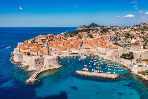 Dubrovniks Altstadt mit Festungsmauer und Hafen, Blick von oben, Foto: DaLiu/shutterstock Das Bild zeigt eine von einer mächtigen Steinmauer umgebene Altstadt an der kroatischen Adriaküste. Die Dächer sind in kräftigem Orange gehalten, dahinter erhebt sich hügeliges, grünes Umland. Im Vordergrund liegt ein kleiner Hafen mit weißen Booten im türkisblauen Wasser. Eine Festung ragt an der linken Seite ins Meer, über dem Wasser fliegt eine Möwe.