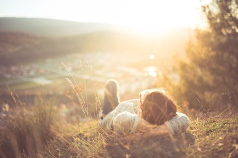 Foto: eldar nurkovic / shutterstock Eine Frau mit kurzen Haaren und Brille liegt rücklings im Gras auf einem Hügel im Licht der tiefstehenden Sonne. Sie hat die Arme unter dem Kopf verschränkt und schaut entspannt in den Himmel. Sie ist dabei nur von hinten zu sehen.