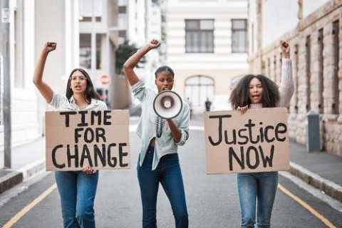 Das Bild zeigt drei Personen, die mitten auf einer Straße stehen und jeweils ein Schild halten. Die Person auf der linken Seite hält ein Schild mit der Aufschrift "ZEIT FÜR VERÄNDERUNG", während die Person auf der rechten Seite ein Schild mit der Aufschrift "Gerechtigkeit JETZT" hält. Die Person in der Mitte hält ein Megafon in der Hand und hat den Arm erhoben. Die Gesichter aller drei Personen sind unkenntlich gemacht. Sie stehen dicht beieinander und nehmen die Mitte der Straße ein, die zu beiden Seiten von Gebäuden gesäumt ist. Die Straße ist mit einer gelben Linie in der Mitte markiert, und die Gebäude im Hintergrund haben mehrere Fenster und eine Mischung aus architektonischen Stilen. 