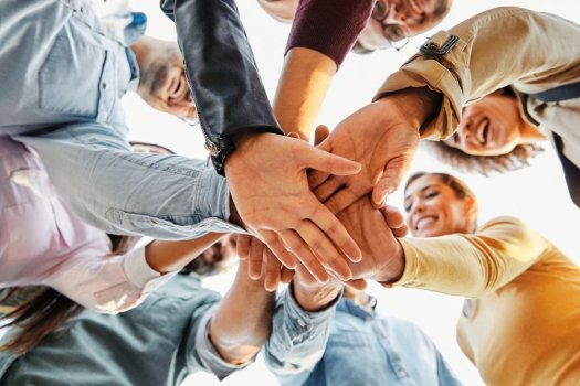 Eine Gruppe von acht Menschen steht im Kreis und streckt von oben betrachtet ihre Hände in die Mitte. Alle Hände berühren sich und bilden einen gemeinsamen Mittelpunkt. Die Personen lachen oder lächeln, sie wirken entspannt und verbunden. Das Bild ist hell belichtet, die Perspektive kommt von unten nach oben. Es vermittelt Zusammenhalt und Vertrauen.