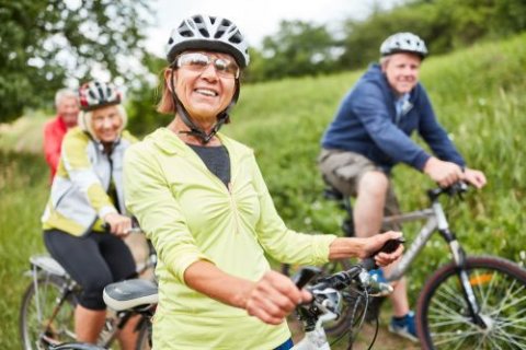 Strahlende Aussichten, Foto: Kneschke/shutterstock Vier ältere Menschen fahren auf Fahrrädern einen Wiesenweg entlang. Im Vordergrund steht eine Frau mit gelber Jacke, Helm und Sonnenbrille, die lächelnd in die Kamera schaut. Hinter ihr folgen zwei Frauen und ein Mann, ebenfalls mit Helmen. Alle wirken fröhlich und sportlich aktiv. Die Umgebung ist grün und sommerlich, mit Bäumen und Wiese im Hintergrund.