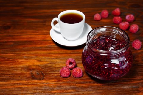 Auf einem Holztisch stehen in der rechten Bildhälfte ein Glas roter Marmelade, eine weiße Tasse mit Untertasse mit einem rötlichen Tee. Drumherum liegen Himbeeren. 