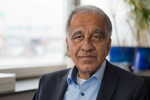 Ein älterer Mann mit grauen Haaren und ernster Miene sitzt in einem Büro. Er trägt ein hellblaues Hemd mit dunklem Sakko. Im Hintergrund sind unscharf Fenster, Pflanzen und Bildschirme zu erkennen.