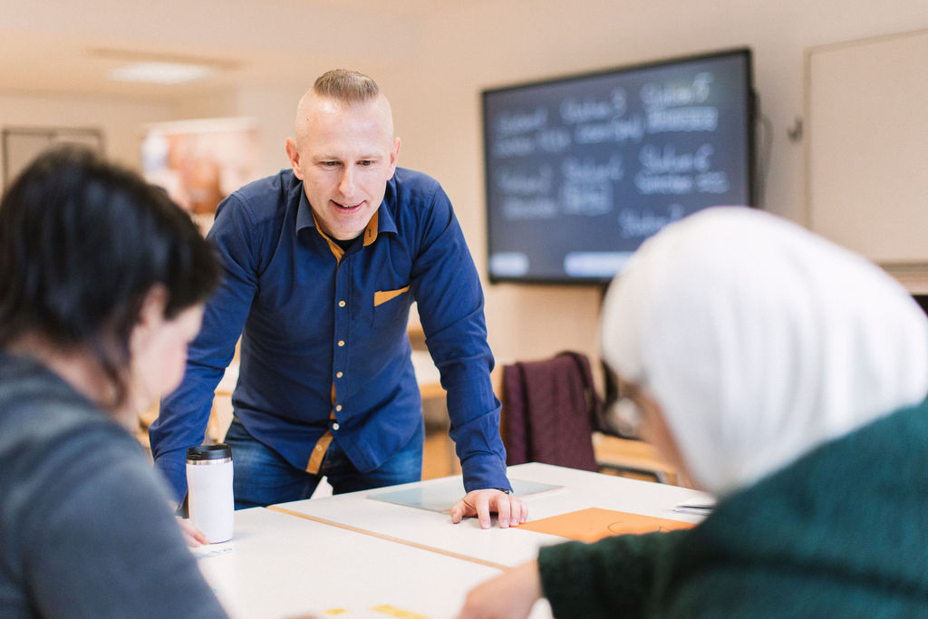 as Bild zeigt ein Klassenzimmer oder eine Besprechung mit drei Personen. Die zentrale Figur steht und beugt sich nach vorne, beide Hände auf einen Tisch gelegt. Diese Person trägt ein blaues Hemd mit orangefarbenen Akzenten und eine blaue Jeans. Links sitzt eine weitere Person, teilweise sichtbar, mit dunklen Haaren und dunklem Oberteil. Vor dieser Person liegt ein weißer Reisebecher. Rechts sitzt eine dritte Person mit weißem Kopftuch. Hinter der stehenden Person befindet sich ein großer Bildschirm mit Text, der eine Liste von Studentennamen oder -gruppen zu sein scheint. Der Raum hat ein modernes Aussehen mit Tischen und Stühlen, die für eine Gruppendiskussion oder Präsentation angeordnet sind.