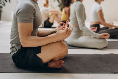 Mehrere Personen sitzen im Schneidersitz auf Yogamatten. Zu sehen sind Beine und Oberkörper, bei den Personen im Vordergrund keine Köpfe. 