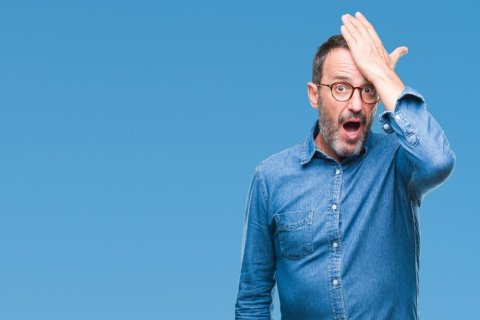 Ein Mann mit grauem Bart, Brille und blauem Jeanshemd schl&auml;gt sich mit der flachen Hand auf die Stirn und rei&szlig;t den Mund auf, als habe er gerade eine Erkenntnis gehabt. Er steht vor einem neutralen blauen Hintergrund. 