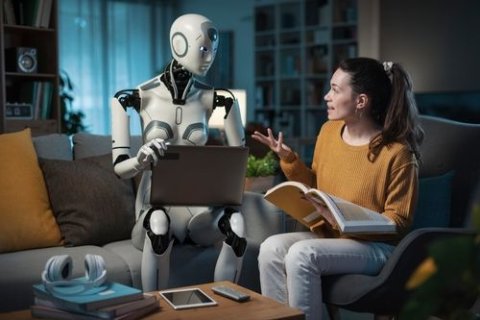 Eine Frau sitzt mit einem Roboter auf einem Sofa und unterh&auml;lt sich. Der Roboter h&auml;lt einen Laptop in der Hand und die Frau ein aufgeschlagenes Buch.