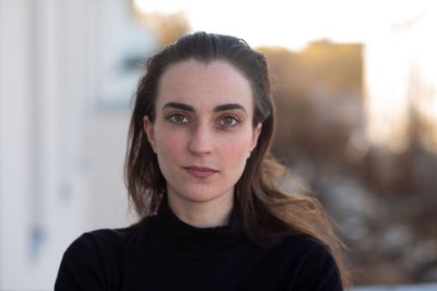 Eine junge Frau mit heller Haut und dunklen, langen Haaren blickt frontal in die Kamera. Sie tr&auml;gt einen schwarzen Rollkragenpullover. Ihr Gesicht ist ruhig, die Lippen geschlossen, die Wangen leicht ger&ouml;tet. Hinter ihr verschwimmt ein heller Hintergrund im Gegenlicht, vermutlich eine Stadt- oder H&auml;userzeile im Abendlicht.