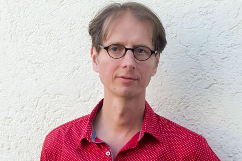 Mann mittleren Alters mit runder, dunkler Brille und hellbraunem, leicht gewelltem Haar vor wei&szlig;er, strukturierter Wand. Blickt direkt in die Kamera, Gesichtsausdruck neutral bis freundlich, Mundwinkel leicht angehoben. Tr&auml;gt rotes Hemd mit kleinen wei&szlig;en Punkten. Bildausschnitt: Kopf und Oberk&ouml;rper bis zur Brust. Tageslichtausleuchtung, frontal zentrierte Aufnahme.