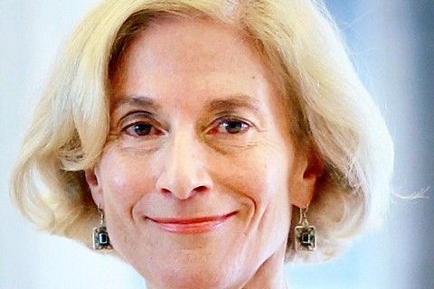 Nahaufnahme einer &auml;lteren Frau mit schulterlangem, wei&szlig;blondem Haar. Sie l&auml;chelt zur&uuml;ckhaltend, der Blick geht leicht zur Seite. Helle Haut, markante Wangenknochen. Sie tr&auml;gt rechteckige Ohrh&auml;nger mit dunkelgr&uuml;nen Steinen in silberner Fassung. Hintergrund unscharf, hellblau-wei&szlig;.