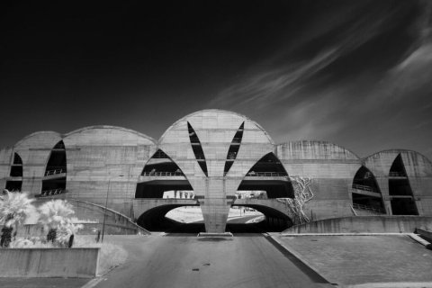 Schwarz-Wei&szlig;-Aufnahme eines brutalistischen Betonbaus: Mehrere halbkreisf&ouml;rmige Schalen aus Sichtbeton reihen sich zu einer symmetrischen Fassade, durchbrochen von schmalen, blattf&ouml;rmigen &Ouml;ffnungen. In der Mitte f&uuml;hrt eine leere Stra&szlig;e unter das Geb&auml;ude. Links helle Palmen, dar&uuml;ber ein fast schwarzer Himmel mit Schleierwolken.