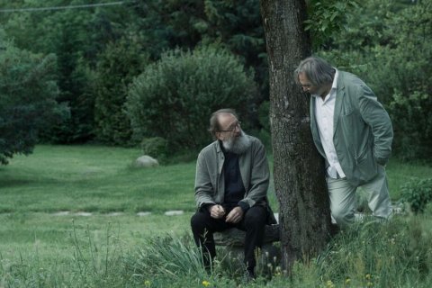 Zwei &auml;ltere M&auml;nner im Gespr&auml;ch in einem sommerlich gr&uuml;nen Garten. Links sitzt Michele De Lucchi mit grauem Vollbart und Brille auf einem Steinblock, er tr&auml;gt eine graugr&uuml;ne Jacke &uuml;ber schwarzer Kleidung. Rechts lehnt Victor Kossakovsky mit grauem Haar an einem Baumstamm, in hellem Anzug und wei&szlig;em Hemd, den Blick zu De Lucchi gewandt. Um sie herum hohes Gras, Str&auml;ucher und B&auml;ume in ged&auml;mpften Gr&uuml;nt&ouml;nen.