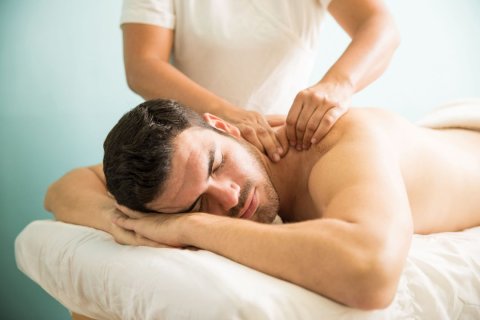 Auf einer Massageliege liegt b&auml;uchlings ein Mann mit geschlossenen Augen und nacktem Oberk&ouml;rper. Wir sehen ihn von der H&uuml;fte aufw&auml;rts. Neben der Liege steht eine Person in wei&szlig;em Shirt, von der wir nur Oberk&ouml;rper und Arme sehen, und massiert ihm den Nacken. 