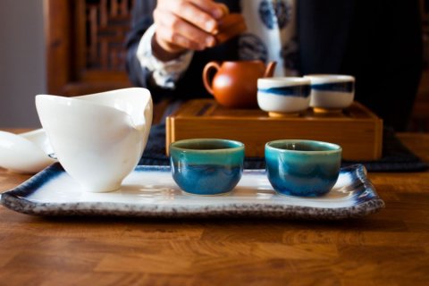 Bild eines chinesischen Teegedecks - bestehend aus einer wei&szlig;en Kanne und zwei kleinen Tassen auf einem Keramiktablett - auf einem Holztisch; im Hintergrund sieht man die Hand einer Person, die den Deckel einer weiteren Keramik-Teekanne &ouml;ffnet, neben der zwei weitere Teetassen stehen.