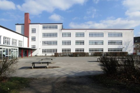 Dreigeschossiges, wei&szlig; verputztes Schulgeb&auml;ude mit breiten Bandfenstern. Markanter roter Schornstein auf dem Dach. Im Vordergrund ein leerer Betonschulhof mit zwei Sitzb&auml;nken und niedrigen Hecken. Links ein Eingangsvorbau mit roten St&uuml;tzen. Blauer Himmel mit vereinzelten Wolken. 