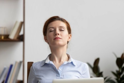 Eine Frau mit Bluse sitzt auf einem Holzstuhl vor - bzw. aus der Betrachterperspektive hinter - einem Laptop-Bildschirm, von dem wir nur den oberen Rand sehen. Sie h&auml;lt den Kopf leicht nach hinten &uuml;berdehnt, streckt dabei den Hals und hat die Augen geschlossen. 