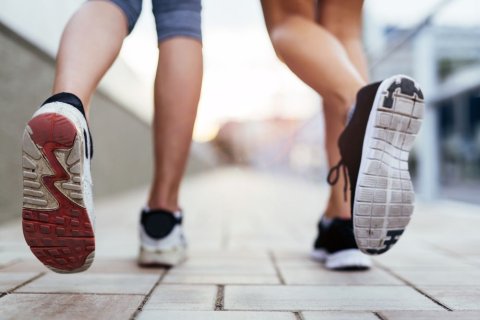 In Nahaufnahme sind die Unterschenkel und F&uuml;&szlig;e zweier Personen beim Laufen auf einem gepflasterten Gehweg zu sehen. Die F&uuml;&szlig;e sind mit Sportschuhen bekleidet, wobei jeweils das linke Bein im Laufen gerade nach hinten, Richtung Kamera, gestreckt ist, so dass die Schuhsohlen zu sehen sind. 
