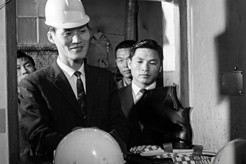 Schwarz-Wei&szlig;-Aufnahme, 1960er Jahre. Im Vordergrund ein Mann mit wei&szlig;em Schutzhelm, Anzug und Krawatte, der leicht l&auml;chelt. Neben ihm ein zweiter Mann im Anzug, ernst blickend. Im Hintergrund weitere M&auml;nner. Auf dem Tresen: ein weiterer Helm, ein schwarzer Stiefel, gefaltetes Stoff. Enger Raumausschnitt, Durchreiche oder Ausgabefenster.