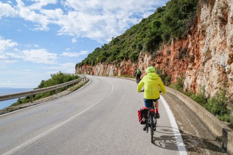 Eine Radfahrerin in gelber Regenjacke mit gelb bezogenem Helm f&auml;hrt auf einer kurvigen K&uuml;stenstra&szlig;e bergauf, am Gep&auml;cktr&auml;ger eine rote Packtasche. Vor ihr zwei weitere Radfahrende. Rechts steigt eine rot-wei&szlig;e Felswand mit gr&uuml;nen B&uuml;schen auf, links begrenzt eine Leitplanke die Stra&szlig;e, dahinter liegt tiefblau das Meer. &Uuml;ber der Szene ein Himmel mit lockeren Wolken.