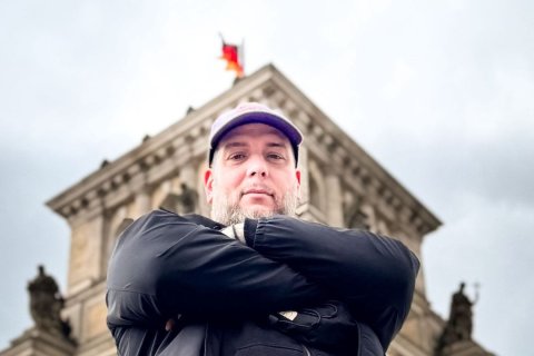 Ein Mann mit grauem Vollbart blickt von oben herab in die Kamera, die Arme vor der Brust verschr&auml;nkt. Er tr&auml;gt eine helle Schirmm&uuml;tze und eine schwarze Jacke. Hinter ihm erhebt sich unscharf der Giebel des Reichstagsgeb&auml;udes in Berlin, auf der Spitze weht eine deutsche Flagge. Auf dem Dach stehen dunkle Statuen. Der Himmel ist grau bew&ouml;lkt, das Licht diffus und k&uuml;hl.