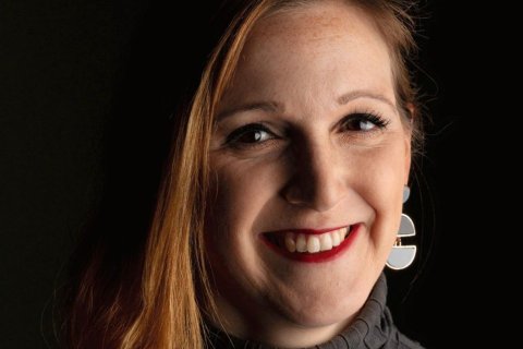 Nahaufnahme des Gesichts einer Frau mit langem rotblondem Haar, das seitlich &uuml;ber die Schulter f&auml;llt. Sie l&auml;chelt breit, tr&auml;gt roten Lippenstift und blickt leicht schr&auml;g in die Kamera. Ihre Augen sind dunkel, auf Nase und Wangen sind Sommersprossen erkennbar. Am linken Ohr h&auml;ngt ein gro&szlig;er Ohrring aus zwei grauen Halbkreisen. Sie tr&auml;gt einen dunkelgrauen Rollkragenpullover. Der Hintergrund ist schwarz, warmes Licht f&auml;llt von der Seite auf ihr Gesicht.
