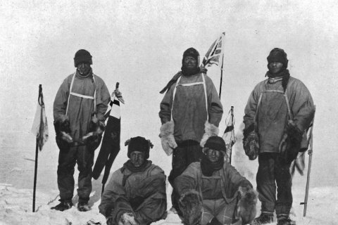 Fünf Männer in dicken Pelzjacken posieren erschöpft im ewigen Eis. Sie stehen und sitzen dicht zusammen, hinter ihnen ragen Fahnen in den Schnee, darunter eine britische Flagge. Ihre Gesichter wirken abgekämpft und eingefroren von der Kälte. Das Schwarz-Weiß-Foto dokumentiert die dramatische Realität der Polarforschung im frühen 20. Jahrhundert und den erbitterten Wettlauf um den Südpol.