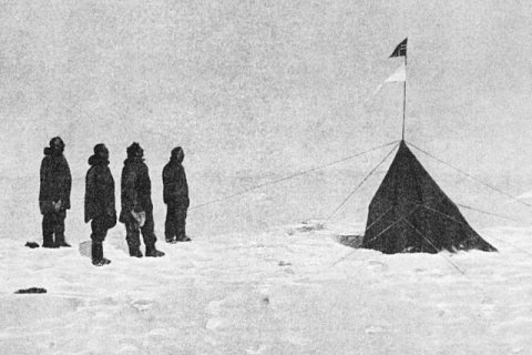 Vier Männer in dicken Polarkleidern stehen in einer Schneelandschaft. Sie blicken auf ein dunkles Zelt, über dem eine Fahnenstange mit der norwegischen Flagge und weiteren Markierungen im Wind flattert. Der Schnee wirkt endlos und leer, der Himmel grau und trostlos. Die Szene zeigt den historischen Moment im Dezember 1911, als Roald Amundsen mit seinem Team als erste Menschen den geographischen Südpol erreichte.