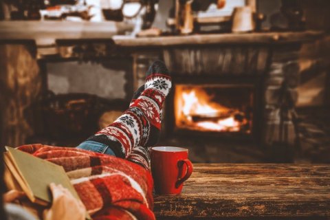 Links vorne im Bild erkennen wir zwei Beine in einer Jeans und mit dicken gestrickten Wollsocken, die zum Teil von einer Wolldecke bedeckt sind. Zwei Hände halten ein Buch. Rechts neben den Beinen steht eine rote Tasse. Die Füße sind auf einer Holzbank abgelegt und im Hintergrund brennt ein Feuer in einem Kamin. Im Foto dominieren warme Farben wie Rot, Orange und Braun. 