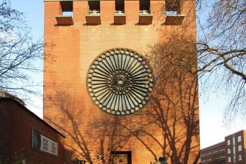 Das Bild zeigt ein großes Backsteingebäude mit einem markanten kreisförmigen architektonischen Merkmal an der Fassade. Dieses kreisförmige Element ist zentral angeordnet und besteht aus einem radialen Muster mit einer Reihe von konzentrischen Kreisen und Speichen, die von der Mitte ausgehen und einem großen Rad oder einer Rosette ähneln. Oberhalb dieses Merkmals befinden sich vier rechteckige Öffnungen oder Fenster mit jeweils einer Kante, die gleichmäßig über den oberen Teil des Gebäudes verteilt sind.
Links vom Gebäude befindet sich ein kleinerer Backsteinbau mit einem Schild mit der Aufschrift "MUSIK KUNST EVENT CHOR FORUM ESSEN". Vor dem Gebäude befindet sich eine Glocke, die auf einem Sockel steht und von niedriger Vegetation umgeben ist. Das Gebäude wird auf beiden Seiten von Bäumen flankiert, in den Rahmen ragen Äste. Im Hintergrund ist rechts ein weiteres Backsteingebäude zu sehen, das Teil eines größeren Komplexes oder Viertels zu sein scheint.