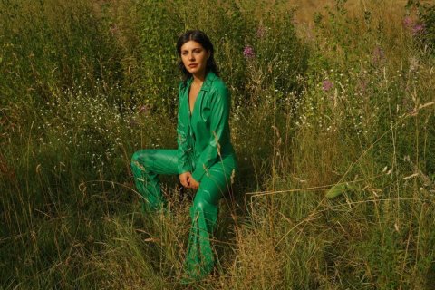 Eine Frau mit dunklem, kinnlangem Haar sitzt auf einer sommerlichen Wiese. Sie trägt einen glänzenden, smaragdgrünen Hosenanzug mit tiefem V-Ausschnitt. Ihre Hände ruhen locker auf den Oberschenkeln, der Blick geht direkt in die Kamera. Das Gras reicht ihr bis zur Hüfte, dazwischen wachsen weiße und violette Wildblumen. Warmes, goldenes Licht fällt von links auf die Szene.