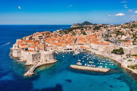 Dubrovniks Altstadt mit Festungsmauer und Hafen, Blick von oben, Foto: DaLiu/shutterstock Das Bild zeigt eine von einer mächtigen Steinmauer umgebene Altstadt an der kroatischen Adriaküste. Die Dächer sind in kräftigem Orange gehalten, dahinter erhebt sich hügeliges, grünes Umland. Im Vordergrund liegt ein kleiner Hafen mit weißen Booten im türkisblauen Wasser. Eine Festung ragt an der linken Seite ins Meer, über dem Wasser fliegt eine Möwe.